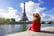 Young traveler woman in red dress and hat sitting on the quay of Seine River looking at Eiffel Tower