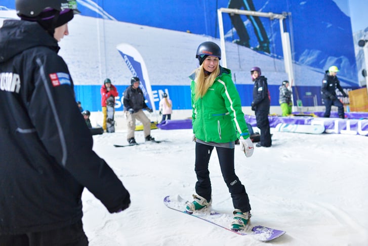 A woman in a green coat receeiving snowboarding lessons