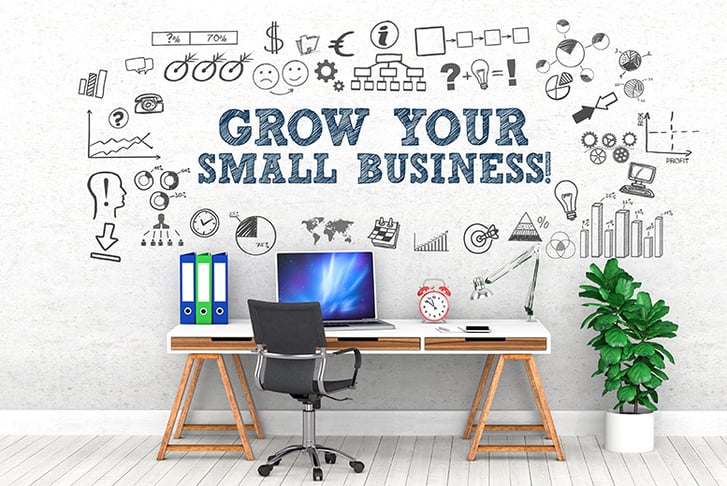 A desk with a laptop open on it against a wall that has 'Grow Your Small Business' written on it with diagrams and doodles surronding